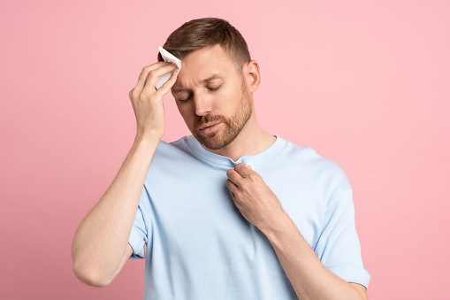 Man suffering from heat stuffiness wiping sweat on forehead with paper napkin isolated on pink background. Exhausted overheated bearded male with closed eyes. Summer hot weather, stuffy room concept.