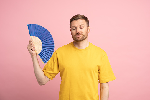 Overheated man waving paper fan suffer from heat, feels sluggish and drowsiness. Unhappy exhausted guy cooling in hot summer weather isolated on studio pink background. Overheating, stuffiness concept