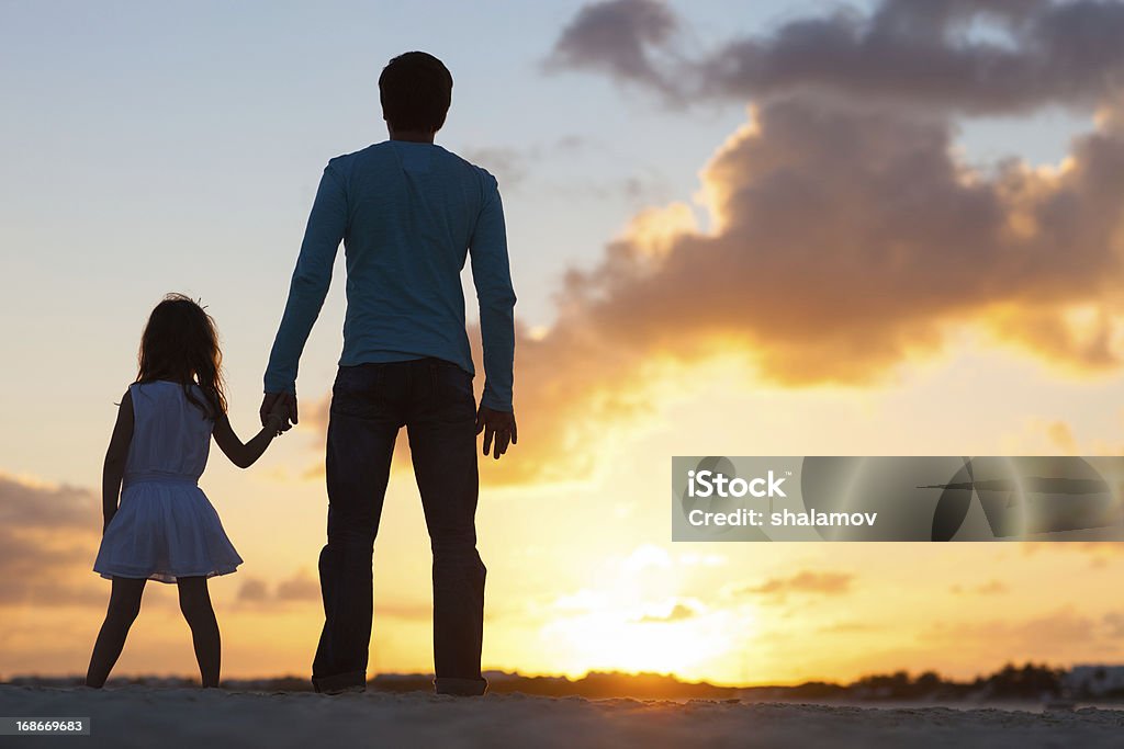 Father and daughter holding hands watching sunset Father and little daughter silhouettes on beach at sunset Adult Stock Photo
