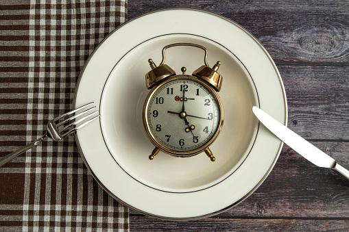 Time to eat. Meal planning for diet concept. Alarm clock, plate, fork knife on a wooden background.