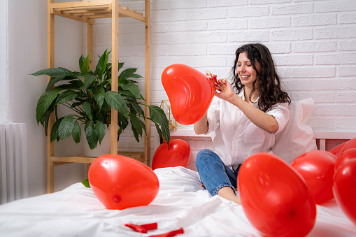 Girl is blowing air balloon at home Handsome brunette girl inflating red heart shaped balloon sitting on bed. Preparation for valentines day party, celebration, dating and surprise.