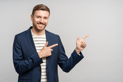 Smiling businessman points with index fingers on empty space for ad on gray studio background. Confident happy middle age promoter man looking at camera showing by forefinger at blank copy space.