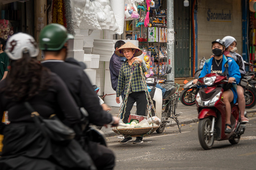 A woman with straw hat carries goods in baskets over her shoulder in Hanoi, Vietnam. Motorcyclists pass her in the street