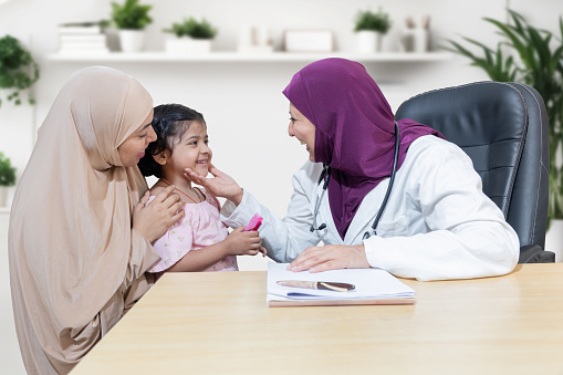 Smiling young muslim doctor wearing hijab checking girl child at modern clinic. Friendly female pediatrician examine young kid patient with her mother.