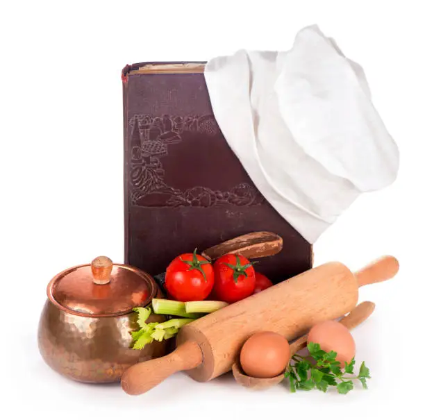 Photo of Still life with a book of ancient recipes. kitchen equipment - copper pan, rolling pin, spoons and different raw vegetables on a white background