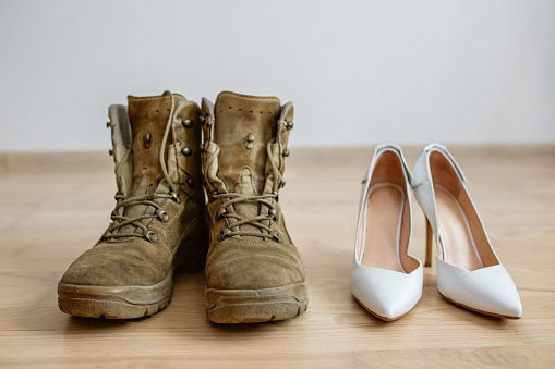 Old worn military boots, women's white shoes on wooden floor. Veteran and family concept. Copy space