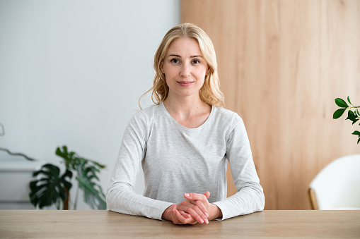 Smiling woman looking at camera, sitting at wooden table in conference room. Listening carefully coach or mentor. Audience at training seminar or online meeting. Business portrait of teacher