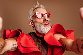 Mature gay man in beautiful red dress making self portrait and blowing a kiss against brown background