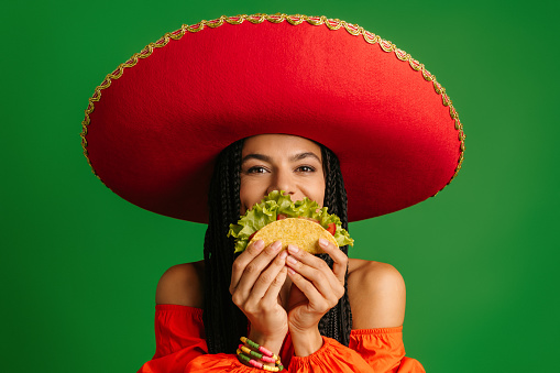 Cheerful Mexican woman in Sombrero hiding half face behind taco against green background
