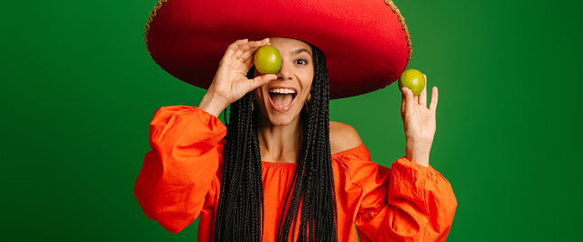 Happy young Mexican woman in Sombrero holding fresh limes and smiling against green background