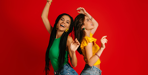 Two attractive young women in colorful wear dancing against red background