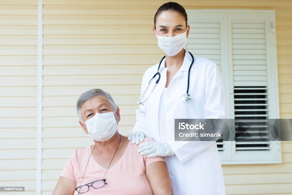 Vaccination Senior woman wearing a protective face mask is getting home visit  from a female doctor with white surgical gloves and protective face mask. They are looking at camera with positive emotion. 70-79 Years Stock Photo