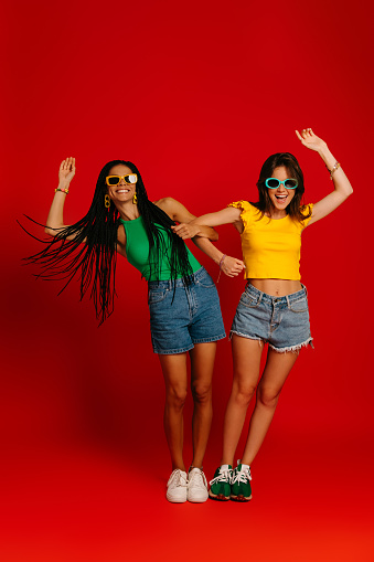 Full length of two playful young women in colorful wear holding hands while having fun against red background
