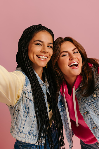 Two playful young women in colorful wear embracing and making self portrait against pink background