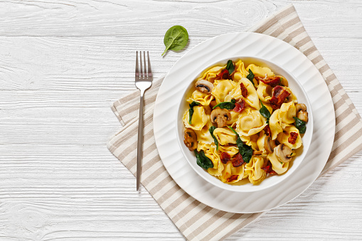 italian tortellini with mushrooms, spinach and fried bacon in white bowl on white wood table, horizontal view from above, flat lay, free space