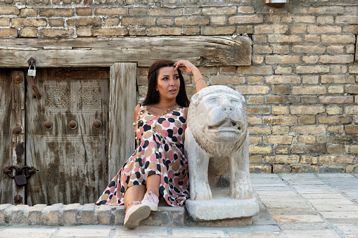 A lady is sitting by the door of a cellar in the fort  of Bukhara, Uzbekistan. Net to her the statue of a lion.