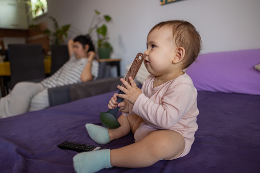 Playful Caucasian 1 year old baby, sitting on the bed and playing with ladle, while her mother sitting near her