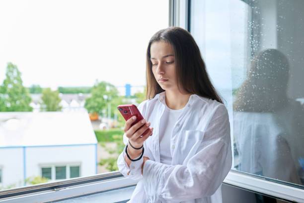 窓の近くの家でスマートフォンを持つ深刻な10代の女の子 - teenager problems typing teenagers only ストックフォトと画像