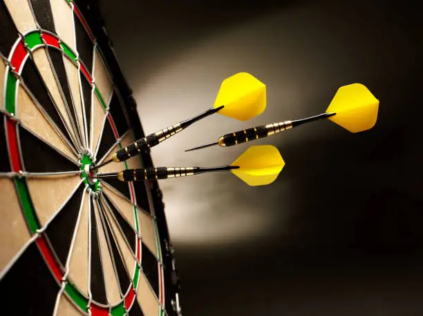 Two yellow darts hitting the target in a game of darts the third is airborne and on target, scoring a bulls eye. 