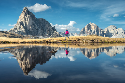 Walking woman and mountains reflected in lake at sunset in autumn in Dolomites, Italy. Standing girl on the shore of lake is looking on high rocks and blue sky with clouds in fall. Reflection in water