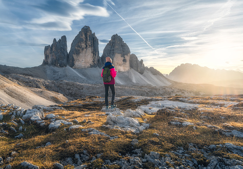 Girl with backpack on the mountain peak and high rocks at colorful sunset in autumn. Tre Cime, Dolomites, Italy. Landscape with young woman on trail, cliffs, sky with clouds and sun in fall. Hiking