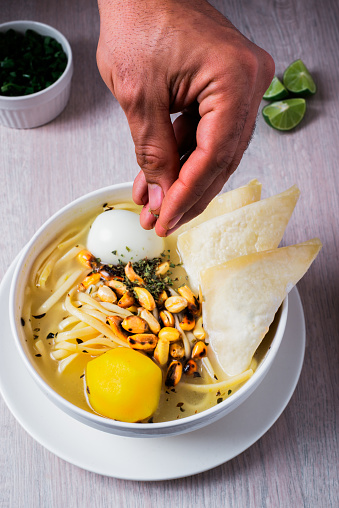 The hand of a Latin man pouring oregano over a chicken soup with wantan, noodles, egg, yellow potato, lemon and Chinese onion.
