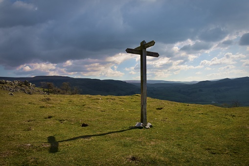 A wooden signpost standing on high grassy moorland casting a shadow on a sunny day with dark hills in the background and cloudy sky