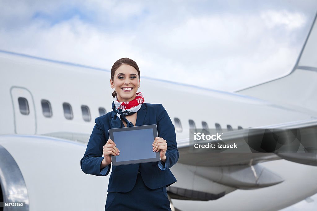 Beautiful air stewardess with a digital tablet Outdoor portrait of a cheerful flight attendant standing in front of an aircraft and showing a digital tablet at the camera. Digital Tablet Stock Photo