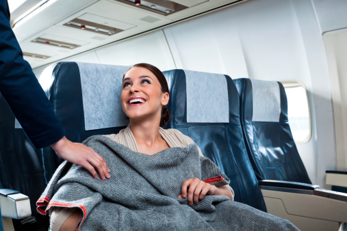 A cheerful young woman sitting on the airplane thanks an air stewardess for a blanket.