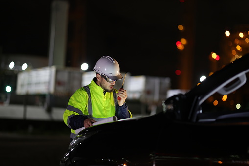 A portrait system engineer with a green safety jacket holds the drawing working on the hood of a car and using a walkie  talkie in a front factory on night shift