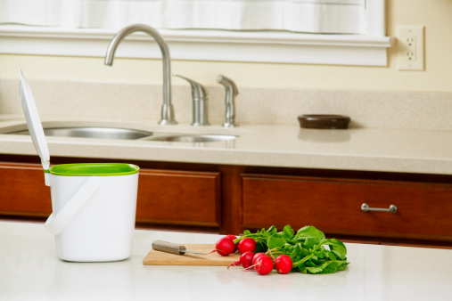 A white kitchen compost bin, with the lid open, sitting on a counter top with a cutting board and fresh radishes. This compost bucket is designed to hold kitchen waste until it is taken to an outdoor compost bin.