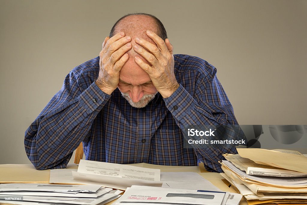 Depressed Senior Adult Man With Stacks of Papers and Envelopes A senior adult man sitting at a table or desk stacked with papers and envelopes looking down with his hands on his head.  Senior Adult Stock Photo