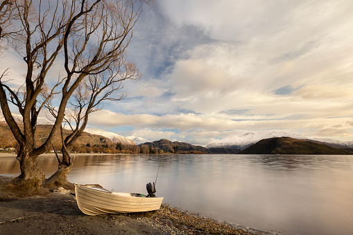 It is early morning at Lake Wanaka, New Zealand, shortly after sunrise. Conditions are calm and a small boat is waiting for it's owner to take it out fishing on the lake. The beach is in Glendhu Bay, a ten minute drive from the town of Wanaka. In the background, you can see the Glendhu Bay campsite that stretches along shore of the lake.