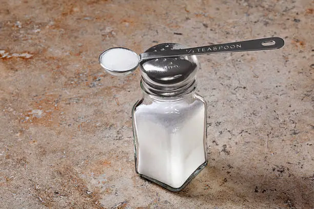 Half teaspoon of salt rests on top of a salt shaker sitting on counter top. The recommended amount of sodium intake is less than one half teaspoon for people over 50 years old, African Americans, individuals with hypertension, diabetes, or chronic kidney disease.