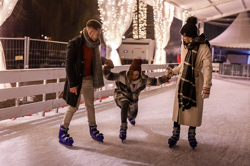 Candid shot of two young people holding their friend's hand while teaching her to ice skate at the ice rink. She is stumbling and falling down.