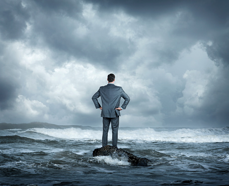 A rear view of a businessman as he stands on a small rock in a rough ocean surf. He stands with his hands on his hips as he looks out across the rough waters and out towards the stormy sea.