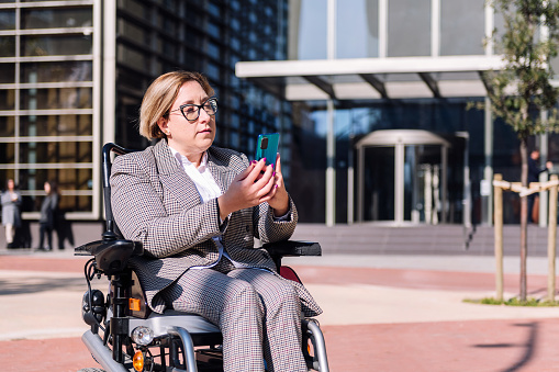 businesswoman in wheelchair using mobile phone in front of an office building in the financial district, concept of diversity and technology of communication