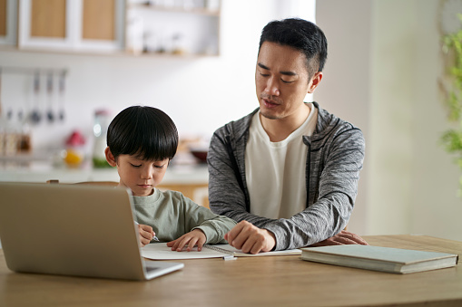young asian father sitting at table tutoring son at home