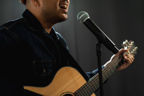 istock Attractive singer man singing music and playing guitar on stage with spotlight strike through the darkness at concert event. Musician performing live with light and smoke at hall. 1687768912
