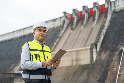 Professional asian maintenance engineer man with safety helmet in construction site dam with hydroelectric power plant and irrigation. Manager engineer man working with tablet at project big building.