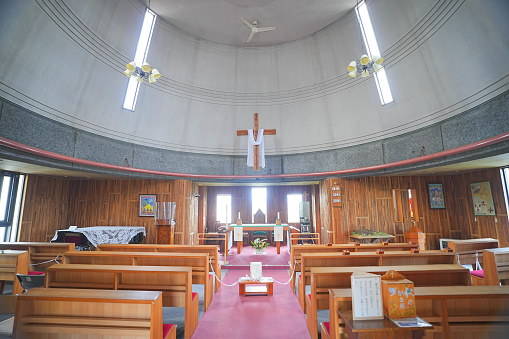 View of the altar of a modern American Catholic Church in Los Angeles. Visible are a crucifix hanging on the wall behind the altar, an ambo (podium) with microphones at right, a celebratory table left of center, an area for musicians in the left toward the back, and seats at the left. The scene is bathed in afternoon sunlight streaming in through windows.