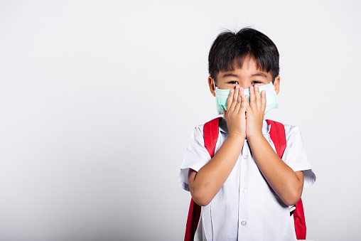 Asian student kid boy wearing student thai uniform and medical protect face mask and hand cover mouth in studio shot isolated on white background, new normal back to school