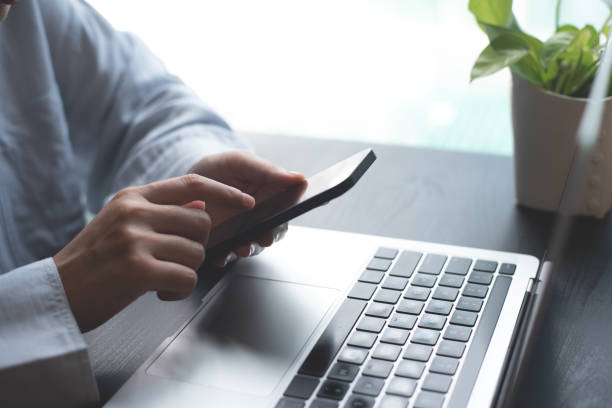 istock Business woman sitting at office table using mobile phone while online working on laptop computer at home office, business casual, telecommuting, working from home, close up 1687900649