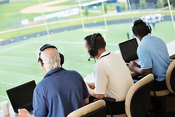 Sportscasters watching and calling a football game in a media press box.