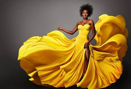 Fashion Woman in Yellow Silk Dress flowing on Wind. Dark Skinned Model with Afro Hairstyle in Long Fantasy Gown over Gray studio background. Happy Stylish Girl dancing