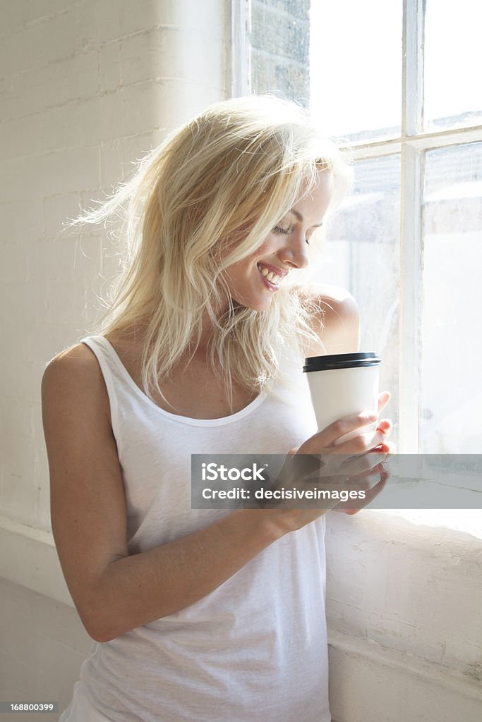 Beautiful woman by window with takeaway coffee A beautiful, happy, young, blonde woman holding a take away beverage standing by a sunny window 20-29 Years Stock Photo