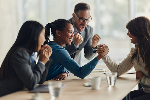 Happy businesswomen competing in arm wrestling in the office while their colleagues are cheering. Focus is on black woman.