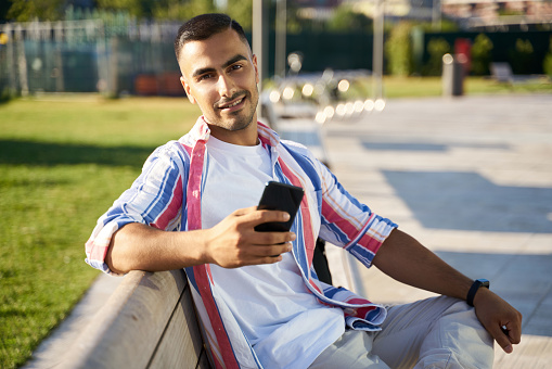 Young smiling handsome middle eastern man using smartphone sitting on bench