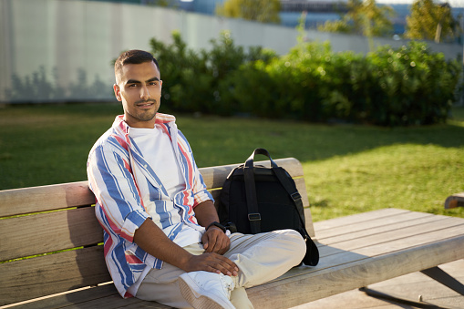 Portrait of smiling handsome middle eastern man with backpack looking at camera sitting on bench outdoors