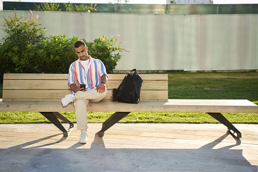 Middle eastern handsome man using mobile phone, communication online sitting on the bench, relaxing outdoors. Young Iranian student sitting in university campus, copy space